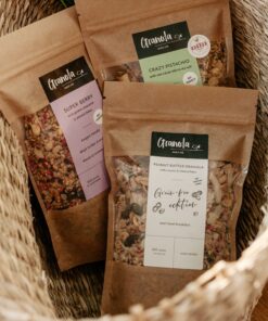 trzy paczki granoli naturalnie bez glutenu w koszyku: granola z owocami liofilizowanymi, granola opiekana w maśle orzechowym bez cukru, granola z pistacjami.