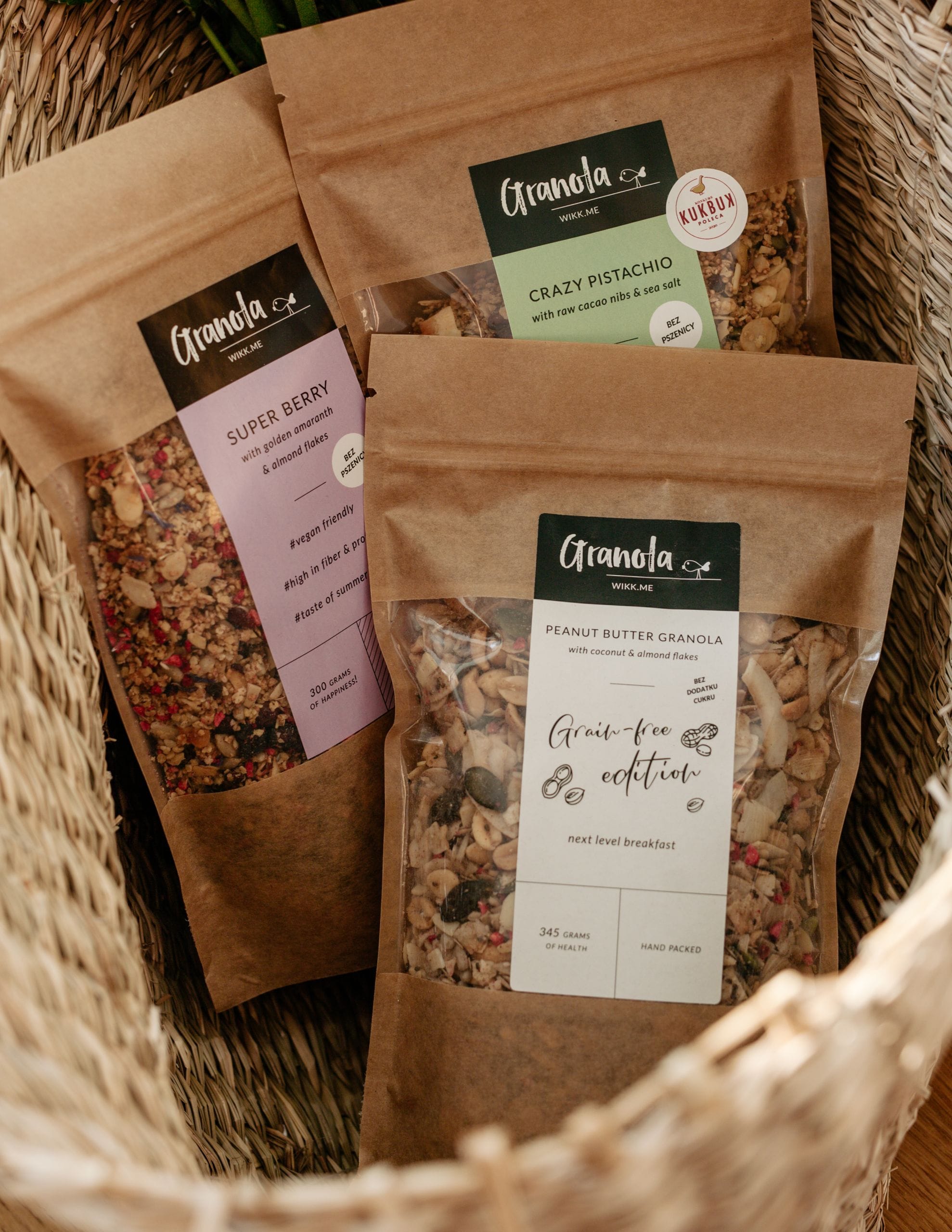 trzy paczki granoli naturalnie bez glutenu w koszyku: granola z owocami liofilizowanymi, granola opiekana w maśle orzechowym bez cukru, granola z pistacjami.